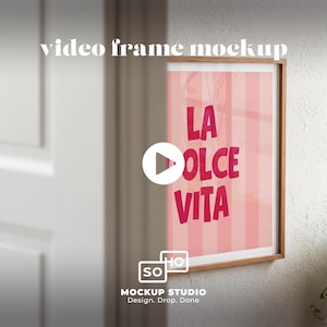 May include: A framed print with the words "LA DOLCE VITA" in pink, set against a pink striped background. The frame is made of wood. The image is a video frame mockup, with the Mockup Studio logo at the bottom.