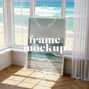 May include: A white framed art print leaning against a window, featuring a watercolor painting of ocean waves. The words "frame mockup" are displayed on the artwork. The window overlooks a beach and ocean.