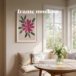 May include: A framed floral art print with a pink flower and green leaves hangs on a beige wall. The words "frame mockup" are at the top. A wooden table with a vase of white flowers sits in front of a window.