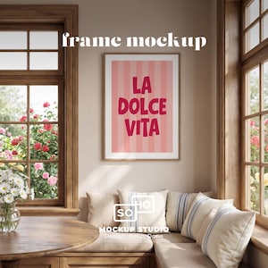 May include: A framed print with the words "LA DOLCE VITA" in a bold, pink font against a striped pink background. The frame is a light brown color. The image is in a room with a window and a built-in bench with pillows.