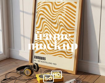May include: A framed poster with a wavy, abstract design in shades of yellow and cream, leaning against a wall. The poster is in a light wood frame. A pair of headphones, a yellow cassette player, and cassettes are on the wooden floor.