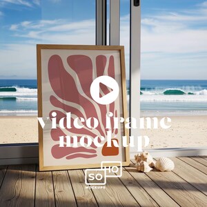 May include: A wooden framed art print with a modern abstract design in shades of pink and orange. The words "video frame mockup" are printed on the art. The frame is propped up against a window overlooking a beach and ocean.