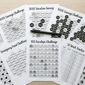 May include: A collection of white paper financial planning sheets with black text and graphics. Titles include "500 Savings Challenge", "1000 Savings Challenge", "3000 Vacation Saving", and "100 Envelope Challenge". A black pen rests on one sheet.