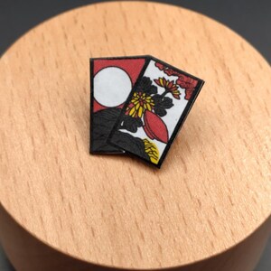 Könnte beinhalten: Zwei kleine, rechteckige Anstecknadeln mit japanischen Kunstwerken. Eine Nadel zeigt einen roten Hintergrund mit einem weißen Kreis und einer schwarzen Bergsilhouette. Die andere Nadel hat einen weißen Hintergrund mit roten, gelben und schwarzen floralen Designs.
