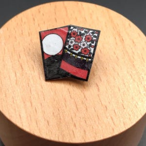 Könnte beinhalten: Eine kleine, rechteckige Anstecknadel mit traditionellem japanischem Design. Die Anstecknadel hat ein rot-schwarzes Farbschema mit floralen und kreisförmigen Mustern. Die Anstecknadel liegt auf einer hellbraunen Holzoberfläche.