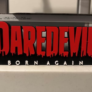 Op de afbeelding: Een 3D-geprint zwart en rood bord met de woorden "Daredevil Born Again" in witte letters. Het woord "Daredevil" is in een druipend rood lettertype.