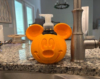May include: Orange Mickey Mouse-shaped soap dispenser with a pump. The dispenser has a carved pumpkin face and two orange ears. The dispenser is sitting on a granite countertop next to a stainless steel faucet.