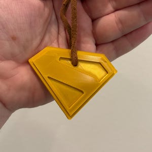 May include: A yellow Superman logo pendant with a brown leather cord. The pendant is diamond-shaped with a raised outline and a textured surface. The cord is attached at the top of the pendant.