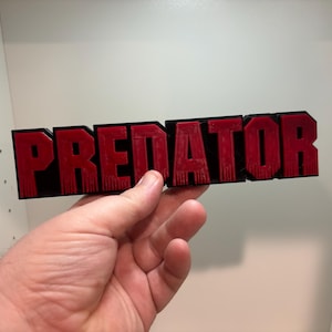 May include: A rectangular sign with the word "PREDATOR" in bold, red letters. The letters are outlined in black. The sign is held in a hand against a white background.