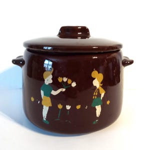 May include: A brown ceramic pot with a lid and two handles. The pot is decorated with painted figures of children and flowers. The figures are wearing green, yellow, and white clothing. The pot is likely vintage or retro.
