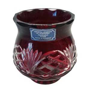 May include: A ruby red crystal vase with a cut glass design. The vase has a scalloped rim and a label that reads "Crystal Legends by Godinger, Handcrafted Crystal, Made in Romania."