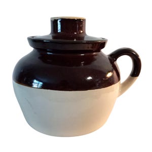 May include: A two-tone ceramic jug with a lid. The upper portion and lid are a glossy dark brown, while the lower portion is off-white. It has a curved handle and a cylindrical neck.