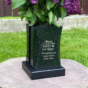 May include: A black granite memorial vase with a floral arrangement of purple lilacs. The vase has the inscription "Always in our hearts, Forever in our Heart" and the name "Jane Swan 1945-2023". The vase is on a stone base.