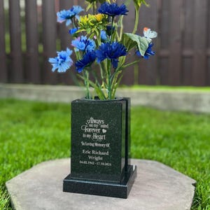 Op de afbeelding: Een zwarte granieten gedenkvaas met een bloemstuk van gele zonnebloemen en blauwe bloemen. De vaas heeft de inscriptie "Always on my mind, Forever in my heart". De vaas staat op een stenen voet.