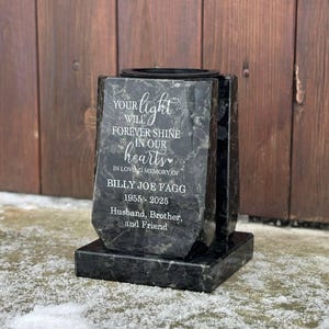 Puede incluir: Una lápida conmemorativa de color gris oscuro con la inscripción "Your light will forever shine in our hearts" y el nombre de Billy Joe Fagg, 1955-2025. El monumento es un homenaje a un esposo, hermano y amigo.