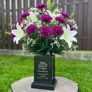 Puede incluir: Un jarrón conmemorativo negro con un arreglo floral de lirios blancos, claveles morados y pequeñas flores blancas. El jarrón tiene la inscripción "Always in my mind, Forever in my heart" y el nombre "Eric Richard Wright".