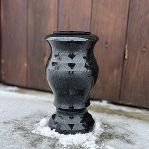 Puede incluir: Bolardo negro de granito con forma decorativa, sobre nieve. El bolardo tiene un acabado texturizado y está colocado frente a una puerta de madera. Altura aproximada: 60 cm.