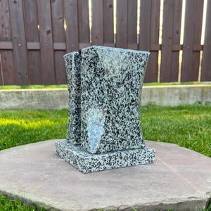 Puede incluir: Una escultura de granito con un patrón moteado en blanco y negro. La escultura tiene una base rectangular y un diseño curvo y abstracto. Se exhibe sobre una piedra grande, plana, de color marrón rojizo, sobre un lecho de hierba verde.