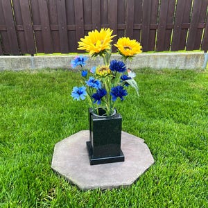Puede incluir: Un jarrón de granito negro con girasoles y flores azules. El jarrón está sobre una base de piedra gris octogonal, colocado sobre un césped verde con una valla de madera al fondo.