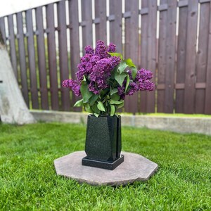 Op de afbeelding: Een zwarte granieten vaas met een boeket paarse seringen. De vaas heeft een geometrisch ontwerp en staat op een vierkante basis, geplaatst op een grote, platte, grijze steen. De achtergrond toont groen gras en een bruine houten schutting.