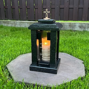Op de afbeelding: Een donkergroene gedenklantaarn met een kruisje bovenop, geplaatst op een steen. Het glazen paneel toont de tekst "You will forever shine in our hearts" en de naam David Wilson. Er staat een brandende kaars in.