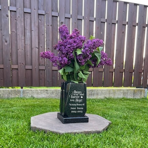 Pode incluir: Um vaso memorial de granito preto com um buquê de flores lilás roxas vibrantes. A superfície frontal está gravada com o texto: Always on my mind, Forever in my Heart, In loving memory of, Jane Swon, 1965-2025, e um pequeno ícone de coração.