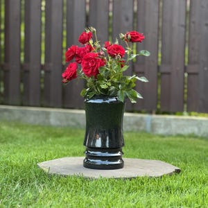 May include: A bouquet of vibrant red roses with green leaves arranged in a glossy black cylindrical vase. The vase features a smooth, reflective surface with subtle speckled details and two distinct horizontal ridges near the base.
