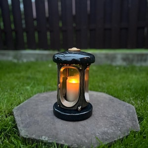 Pode incluir: Uma lanterna verde-escura e dourada com uma vela acesa dentro. A lanterna tem um design decorativo e está sobre uma superfície de pedra, com um fundo desfocado de relva e uma cerca.
