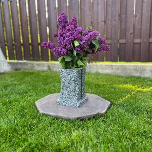 May include: A speckled grey vase, holding a vibrant bouquet of purple lilacs, sits on a large, flat, brown stone. The vase is on a bed of green grass, with a wooden fence in the background.
