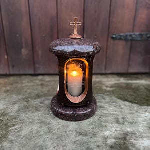 Puede incluir: Una linterna conmemorativa con una cruz en la parte superior, que contiene una vela encendida. La linterna tiene un acabado oscuro y moteado y un marco de color cobre alrededor del cristal. El texto "Your light" y "David Wilson 1964-2024" es visible.
