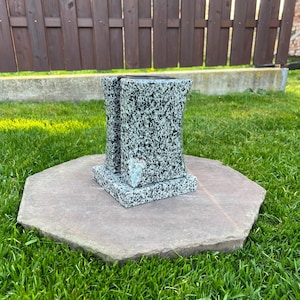 Puede incluir: Un monumento de granito con un patrón moteado gris y negro. El monumento está compuesto por dos pilares y se asienta sobre una base de piedra hexagonal. La base descansa sobre hierba verde, con una valla de madera al fondo.