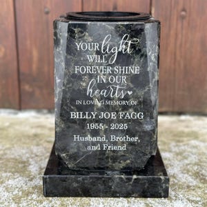 Puede incluir: Urna conmemorativa de mármol negro con la inscripción "YOUR light WILL FOREVER SHINE IN OUR hearts IN LOVING MEMORY OF BILLY JOE FAGG 1955-2025 Husband, Brother, and Friend". La urna tiene una base rectangular y un cuerpo facetado.