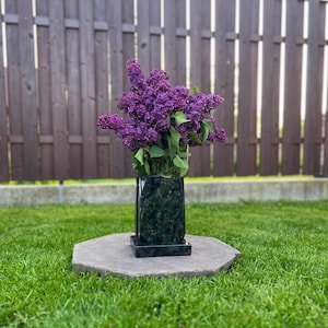 May include: A bouquet of vibrant purple lilacs in a clear glass vase, set within a black marble vase. The vase sits on a gray stone, with a wooden fence and green grass in the background. The flowers are in full bloom.