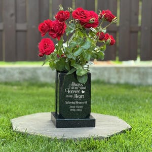 Pode incluir: Um vaso memorial de granito preto com base quadrada e texto gravado em branco que diz: Always on my mind, Forever in my heart, In loving memory of, Jane Swon, 1965-2025. O vaso está preenchido com um buquê de rosas vermelhas e folhas verdes.