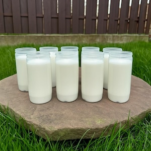 May include: Eight clear plastic cups filled with a white liquid, possibly milk, are arranged on a large, flat, brown stone. The cups are cylindrical with a slightly flared rim. The background shows green grass and a brown wooden fence.