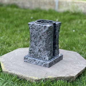Pode incluir: Um vaso funerário retangular de granito com acabamento polido salpicado de azul e cinza. O vaso apresenta uma face frontal côncava e um inserto circular de metal preto no topo, projetado para acomodar arranjos de flores em cemitérios.