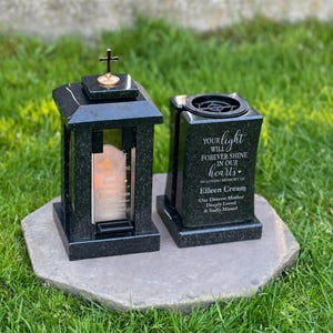 Puede incluir: Dos faroles conmemorativos de granito negro con superficies pulidas. El farol izquierdo presenta una pequeña cruz dorada en la parte superior y un panel de vidrio con el texto 'Your light will forever shine in our hearts, In loving memory of Eileen Crean'.