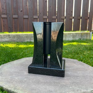Puede incluir: Un jarrón de granito negro con un diseño moderno. El jarrón tiene una base rectangular y dos lados curvos y pulidos que enmarcan una abertura central cilíndrica. El jarrón está sobre una superficie de piedra gris, con una valla de madera y hierba verde al fondo.