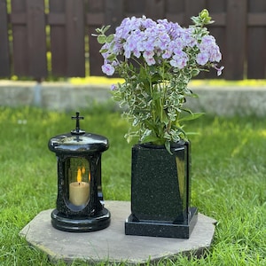 Pode incluir: Uma lanterna funerária de granito preto com uma cruz no topo e uma vela acesa no interior, posicionada ao lado de um vaso retangular de granito preto preenchido com um buquê de flores phlox lilás claro e branco com folhas verdes e brancas.