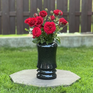 Könnte beinhalten: Eine schwarze Granitvase mit polierter, gesprenkelter Oberfläche und einem gestuften Sockel. Die Vase ist mit einem Strauß leuchtend roter Rosen gefüllt, die mehrere voll erblühte Blumen und einige kleine, geschlossene grüne Knospen zeigen.