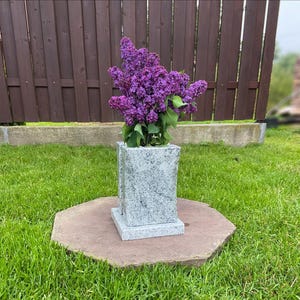 Puede incluir: Un jarrón de granito de forma cuadrada contiene un vibrante ramo de lilas moradas. El jarrón se encuentra sobre una base de piedra octogonal marrón, rodeado de hierba verde. Una valla de madera está en el fondo.
