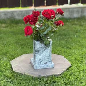Könnte beinhalten: Eine rechteckige Granitvase mit gesprenkelter hellgrauer und schwarzer Oberfläche, die über einen flachen Boden und eine vertikale Öffnung verfügt. Die Vase ist mit einem Strauß leuchtend roter Rosen mit grünen Blättern und Knospen gefüllt.