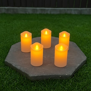 Puede incluir: Cinco velas LED sin llama que funcionan con pilas, con un brillo ámbar cálido. Las velas están dispuestas sobre una piedra hexagonal gris oscuro, sobre un fondo de hierba verde. Cada vela tiene un efecto de parpadeo realista.