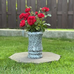 May include: A bouquet of vibrant red roses with green leaves arranged in a speckled, light gray and black cylindrical vase. The vase features a slightly flared top and base with a subtle horizontal ridge detail around the middle section.