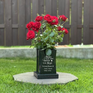 Puede incluir: Un florero conmemorativo de granito negro con base rectangular y cuerpo vertical. El frente está grabado con el texto: Always on my mind Forever in my Heart, In loving Memory of Jane Swon, 1905-2025. Un ramo de rosas rojas se encuentra dentro.