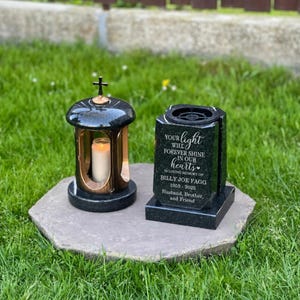 May include: A black granite memorial lantern with a cross topper and a lit candle inside, positioned next to a black granite memorial stone engraved with the text: Your light will forever shine in our hearts, In loving memory of Billy Joe Fagg, 1955-2025, Husband, Brother, and Friend.