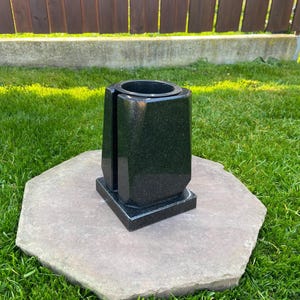 Könnte beinhalten: Eine schwarze Granitvase mit geometrischem Design. Die Vase hat eine zylindrische Öffnung oben und steht auf einem quadratischen Sockel. Sie steht auf einem großen, achteckigen Stein in einer Rasenfläche.