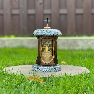 Pode incluir: Uma lanterna memorial com topo e base em padrão de granito, corpo cor de bronze e uma pequena cruz preta no topo. O vidro está gravado com o texto: Your light will forever shine in our hearts, In loving memory of David Wilson 1964-2024.