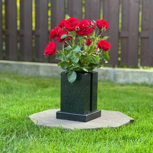 Pode incluir: Um vaso retangular de granito preto com acabamento polido e um detalhe de fenda vertical, contendo um buquê vibrante de rosas vermelho-escuras com folhas verdes. O vaso assenta sobre uma base de pedra plana, com um design sólido e estruturado.