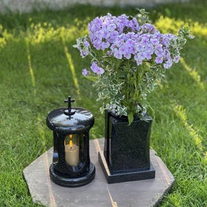 Könnte beinhalten: Eine Grablaterne aus schwarzem Granit mit einem kleinen Kreuz auf der Oberseite und einer brennenden Kerze im Inneren, neben einer quadratischen Vase aus schwarzem Granit mit einem Strauß hellvioletter Phloxblüten und panaschierten Blättern.
